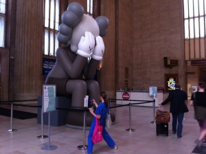A person in blue leans back to take a photo of COMPANION art installation by KAWS at 30th Street Station